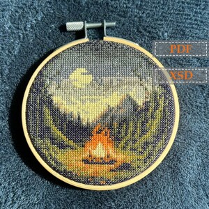 May include: A close-up of a circular embroidery hoop featuring a cross-stitch design of a campfire in a forest beneath a crescent moon. The design is worked in shades of green, brown, orange, and yellow.