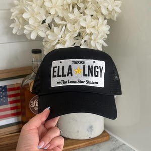 Può includere: Un cappellino trucker nero con una toppa rettangolare bianca sul davanti. La toppa riporta la scritta TEXAS, ELLA, LNGY e The Lone Star State, con una stella gialla posizionata tra ELLA e LNGY, che imita una targa del Texas.