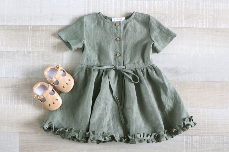 May include: A sage green linen dress with short sleeves and a ruffled hem. The dress has a button closure at the front and a tie at the waist.