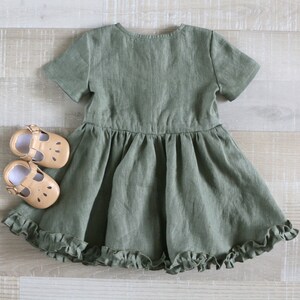 May include: A green linen dress with short sleeves and a ruffled hem. The dress is on a light wood background with two pairs of tan leather shoes.