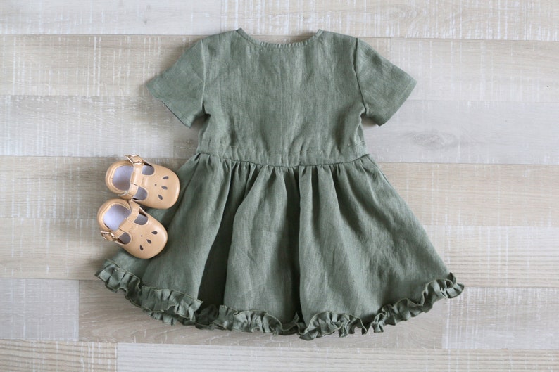 May include: A green linen dress with short sleeves and a ruffled hem. The dress is on a light wood background with two pairs of tan leather shoes.