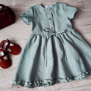 May include: A light green linen dress with a button-up front, a tie at the waist, and a ruffled hem. The dress is photographed on a wooden surface with a small brown purse and red shoes.