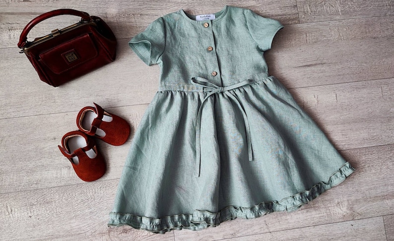 May include: A light green linen dress with a button-up front, a tie at the waist, and a ruffled hem. The dress is photographed on a wooden surface with a small brown purse and red shoes.