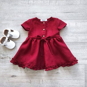 May include: A burgundy linen dress with short sleeves, a button-up front, and a ruffled hem. The dress has a bow tie at the waist.