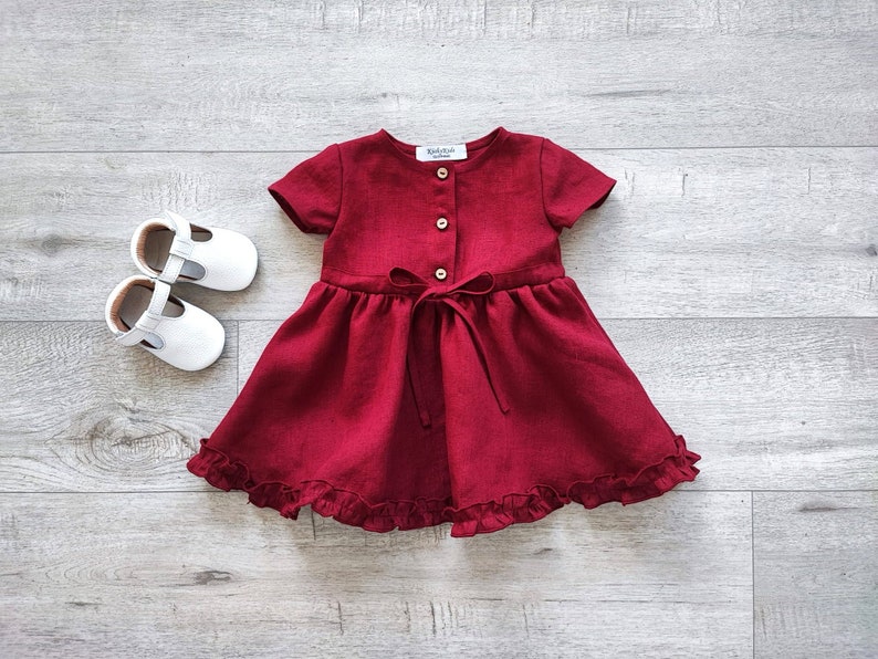 May include: A burgundy linen dress with short sleeves, a button-up front, and a ruffled hem. The dress has a bow tie at the waist.