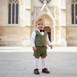May include: A toddler outfit consisting of a white long-sleeved button-down shirt, an olive green double-breasted vest with matching shorts, a dark red bow tie, white knee-high socks, and brown leather shoes, worn by a young child.