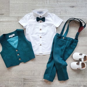 May include: A formal outfit for a baby or toddler, including a white short-sleeved shirt, teal vest, teal pants with suspenders, a teal bow tie, and white shoes. The outfit is laid out on a light wood-grain surface.