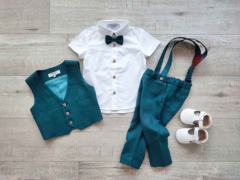 May include: A formal outfit for a baby or toddler, including a white short-sleeved shirt, teal vest, teal pants with suspenders, a teal bow tie, and white shoes. The outfit is laid out on a light wood-grain surface.