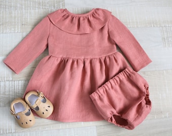 May include: A dusty rose-colored baby dress with long sleeves, a ruffled collar, and a gathered skirt. Matching bloomers and a pair of tan leather baby shoes with cut-out details are also included. The set is made of a soft, textured fabric.