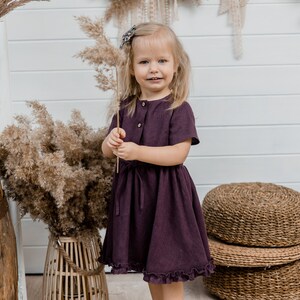 May include: A young girl wearing a dark purple dress with a ruffled hem stands in front of a white wall. She is holding a dried grass stalk in her hand and smiling at the camera.