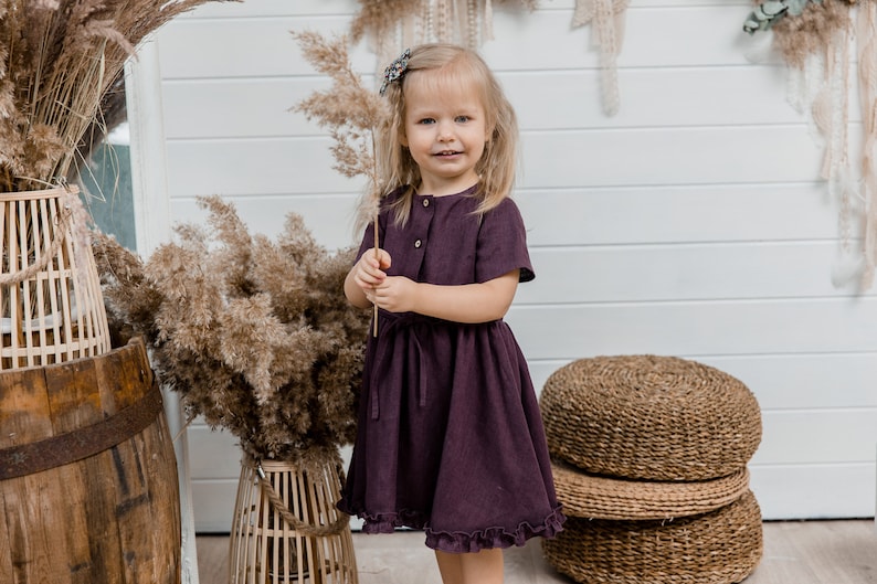 May include: A young girl wearing a dark purple dress with a ruffled hem stands in front of a white wall. She is holding a dried grass stalk in her hand and smiling at the camera.