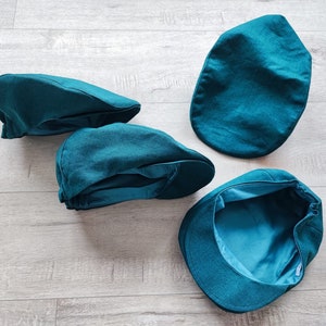 Puede incluir: Cuatro gorras de estilo newsboy de color teal se muestran sobre una superficie clara con vetas de madera. Las gorras, hechas de tela suave, tienen una corona redondeada y una visera pequeña y rígida. El forro interior de una gorra es visible.