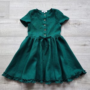 May include: A green linen dress with short sleeves, a button-up front, and a ruffled hem.