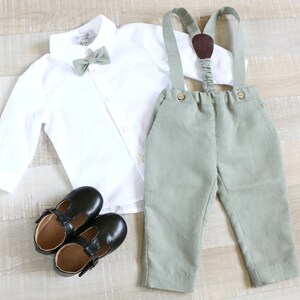 May include: A formal outfit for a child, featuring a white button-down shirt, sage green bow tie, matching suspenders, and pants. Black leather Mary Jane shoes complete the ensemble. The outfit is laid out on a light wood surface.