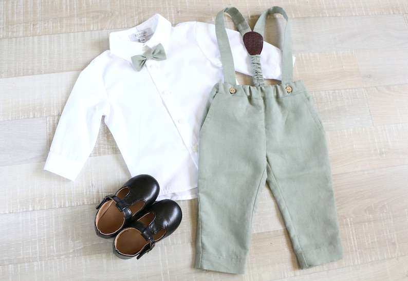 May include: A formal outfit for a child, featuring a white button-down shirt, sage green bow tie, matching suspenders, and pants. Black leather Mary Jane shoes complete the ensemble. The outfit is laid out on a light wood surface.