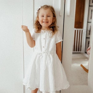 May include: A young girl wearing a white dress with a ruffled hem and a bow at the waist. She is smiling and has her hair pulled back with a white flower clip.