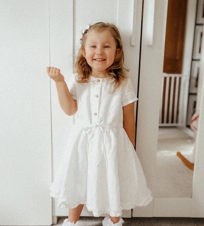May include: A young girl wearing a white dress with a ruffled hem and a bow at the waist. She is smiling and has her hair pulled back with a white flower clip.