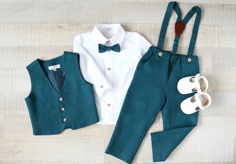 May include: A teal green formal outfit for a child, including a vest, pants with suspenders, a white button-down shirt, a bow tie, and white shoes. The vest and pants have wooden buttons.