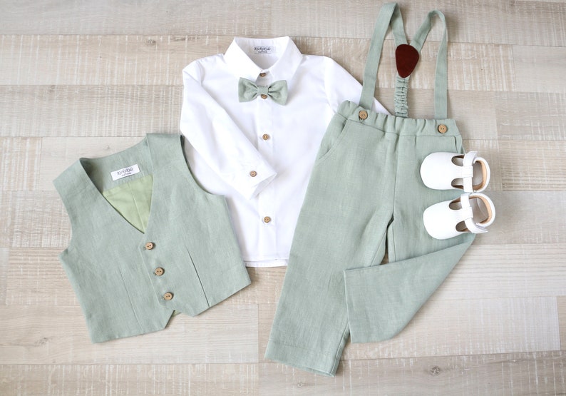 May include: A formal outfit for a baby or toddler, including a white button-down shirt with a bow tie, a green vest, green pants with suspenders, and white shoes. The outfit is in a light green color scheme with wooden buttons.