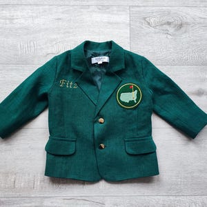Op de afbeelding: Een donkergroen blazer met een gouden knoop en een patch met een golfbaan en de Amerikaanse vlag. De blazer heeft de naam "Fitz" geborduurd op de linkerzijde.