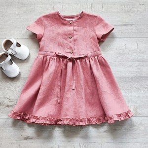 May include: A pink linen dress with short sleeves and a ruffled hem. The dress has a button closure and a tie at the waist.