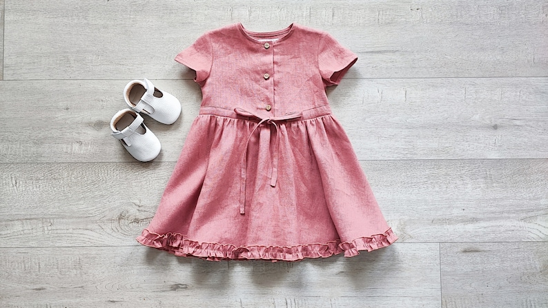 May include: A pink linen dress with short sleeves and a ruffled hem. The dress has a button closure and a tie at the waist.