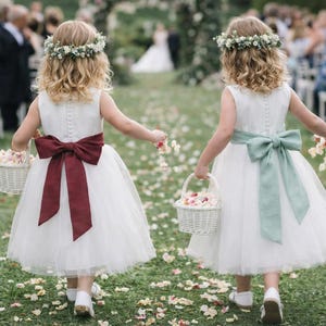 Könnte beinhalten: Zwei Kinder in weißen ärmellosen Kleidern mit Tüllröcken, Blumenkränzen und großen Schleifen in Burgunderrot und Salbeigrün. Jedes Kind trägt einen kleinen weißen geflochtenen Korb voller Blütenblätter auf einem grasbewachsenen Weg.