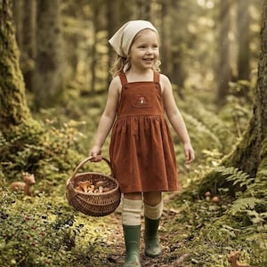 May include: A young child wearing a rust-colored corduroy dress with a mushroom applique, a cream-colored headscarf, and green rain boots. She carries a wicker basket filled with mushrooms while walking through a forest.