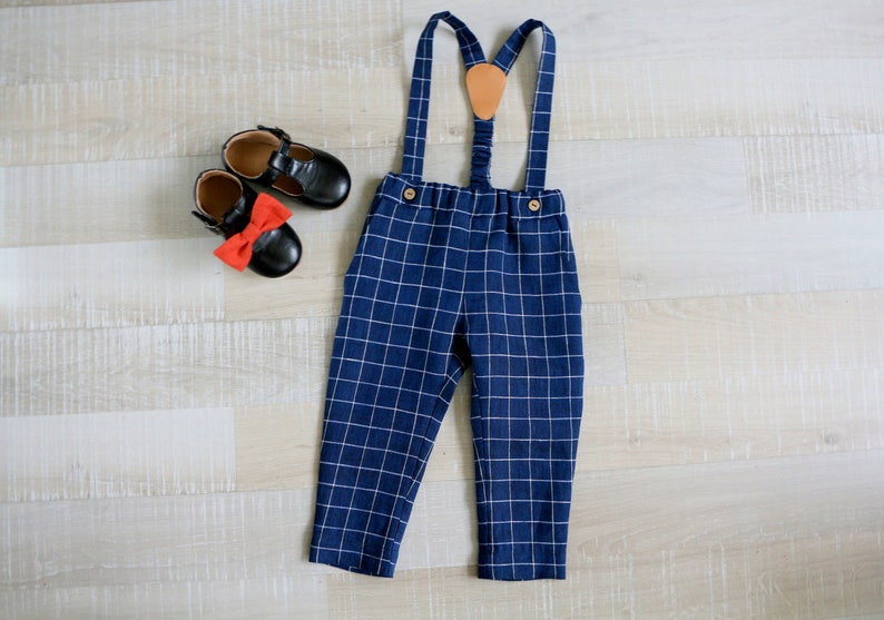 May include: A navy blue plaid suspender pants set with a leather patch and wooden buttons. The outfit is paired with black leather shoes and a red bow. The clothing is designed for children.