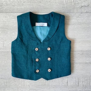 Könnte beinhalten: Eine tealfarbene Weste aus Leinen mit doppelreihigem Verschluss und vier goldenen Knöpfen. Die Weste hat einen Kragen und ist aus einem strukturierten Stoff gefertigt.