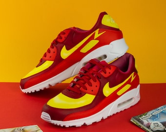 May include: A pair of red and yellow Nike Air Max 90 sneakers with a white sole. The sneakers have a yellow swoosh logo and a yellow lightning bolt design on the side.
