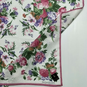 May include: White cotton handkerchief with a pink floral pattern and a pink border. The handkerchief has a small black label with the text "Yves Saint Laurent" in white.