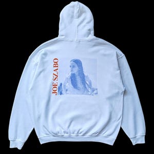 May include: White hooded sweatshirt with a blue and white image of a person on the back. The text "JOE SZABO" is printed vertically in orange on the left side of the sweatshirt.