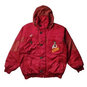 May include: A red hooded jacket featuring a front zip fastening, two chest pockets, and ribbed cuffs. The garment displays a Redskins logo on the left chest, a small logo on the right chest, and textured fabric panels on the sleeves and hood.