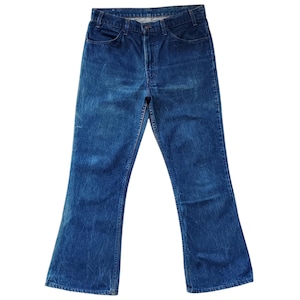 Może przedstawiać: Para dżinsów z niebieskiego denimu w stylu vintage z rozszerzanymi nogawkami. Dżinsy mają klasyczny styl z pięcioma kieszeniami, zapięcie na guzik i zamek błyskawiczny oraz szlufki na pasek. Denim ma wyblakły wygląd.
