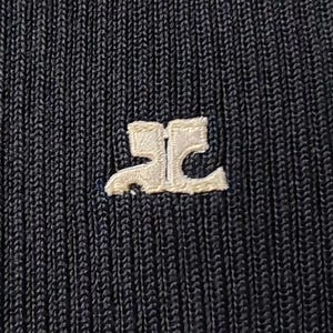 May include: A black ribbed knit fabric featuring a white embroidered logo in the center. The logo consists of two stylized, mirrored letters that form a symmetrical shape. The texture of the fabric is clearly visible with vertical lines.