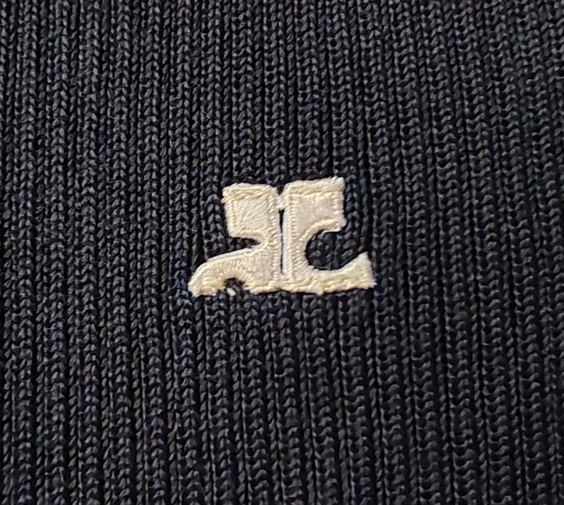 May include: A black ribbed knit fabric featuring a white embroidered logo in the center. The logo consists of two stylized, mirrored letters that form a symmetrical shape. The texture of the fabric is clearly visible with vertical lines.