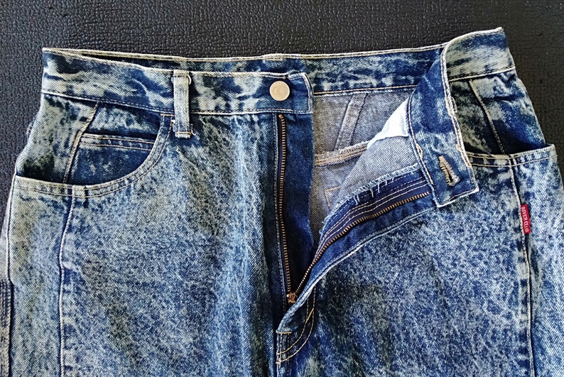 Może przedstawiać: Dżinsy z efektem acid wash z zamkiem błyskawicznym i guzikiem. Dżinsy są jasnoniebieskie z białym wzorem w kropki.