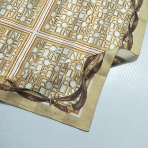 May include: A beige silk scarf with a brown and white pattern. The pattern features the word "CELINE" repeated throughout. The scarf has a brown leather trim with a buckle detail.