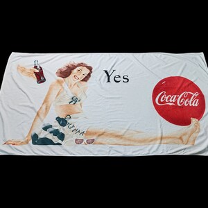 May include: White beach towel with a vintage Coca-Cola advert. The design shows a smiling woman in swimwear, a bottle of Coca-Cola, and the word "Yes". The Coca-Cola logo is in red.