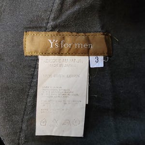 Puede incluir: Primer plano de una etiqueta de prenda con el texto "Y's for men". La etiqueta es marr&oacute;n con texto blanco y una etiqueta blanca con el n&uacute;mero "3". La etiqueta tambi&eacute;n incluye texto en franc&eacute;s e ingl&eacute;s, lo que indica que est&aacute; fabricada en Jap&oacute;n.