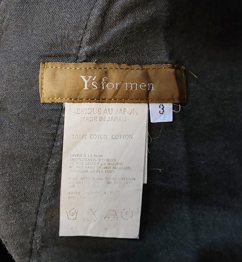 Puede incluir: Primer plano de una etiqueta de prenda con el texto "Y's for men". La etiqueta es marr&oacute;n con texto blanco y una etiqueta blanca con el n&uacute;mero "3". La etiqueta tambi&eacute;n incluye texto en franc&eacute;s e ingl&eacute;s, lo que indica que est&aacute; fabricada en Jap&oacute;n.