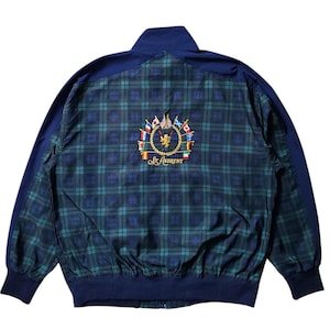 Puede incluir: Chaqueta a cuadros verde y azul marino con cuello y mangas azul marino. La parte trasera de la chaqueta presenta un emblema bordado con las palabras "St. Andrews" y un escudo con banderas.
