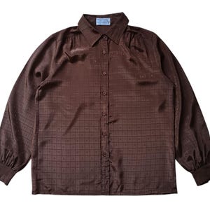 Puede incluir: Camisa marrón de manga larga con cuello y botones. La tela presenta un patrón cuadrado repetido con una 'V' dentro de cada cuadrado. Las mangas están recogidas en los hombros y puños, creando un efecto abullonado.