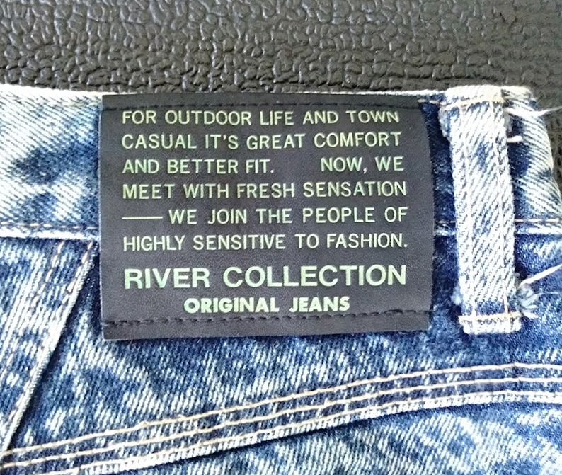 Może przedstawiać: Czarna sk&oacute;rzana naszywka z białym tekstem na niebieskich dżinsach. Tekst brzmi: "FOR OUTDOOR LIFE AND TOWN CASUAL IT'S GREAT COMFORT AND BETTER FIT. NOW, WE MEET WITH FRESH SENSATION -WE JOIN THE PEOPLE OF HIGHLY SENSITIVE TO FASHION. RIVER COLLECTION ORIGINAL JEANS"