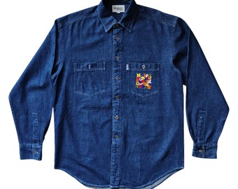 Puede incluir: Camisa vaquera de manga larga en un lavado azul medio. La camisa tiene dos bolsillos en el pecho y una botonadura frontal. Un parche bordado colorido está en el bolsillo izquierdo del pecho. Cuello clásico y puños abotonados.