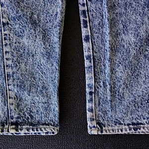 Może przedstawiać: Zbliżenie na dolne brzegi pary niebieskich jeans&oacute;w z efektem acid wash. Jeansy mają wyblakły, przetarty wygląd.
