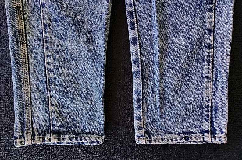 Może przedstawiać: Zbliżenie na dolne brzegi pary niebieskich jeans&oacute;w z efektem acid wash. Jeansy mają wyblakły, przetarty wygląd.