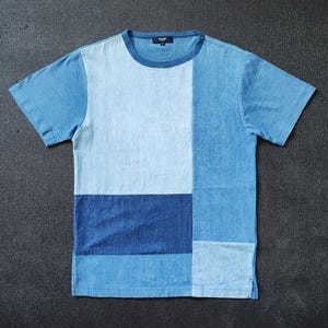 May include: A blue patchwork t-shirt with a crew neck and short sleeves. The shirt features a colour block design with various shades of blue, including light blue, medium blue, and dark blue. The brand name is visible on the collar.