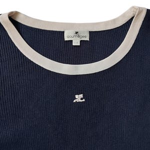 May include: A dark navy blue ribbed knit top featuring a round neckline with a contrasting cream-colored trim. The garment includes a cream-colored embroidered logo on the chest and a white interior label with the text Courr&egrave;ges printed on it.
